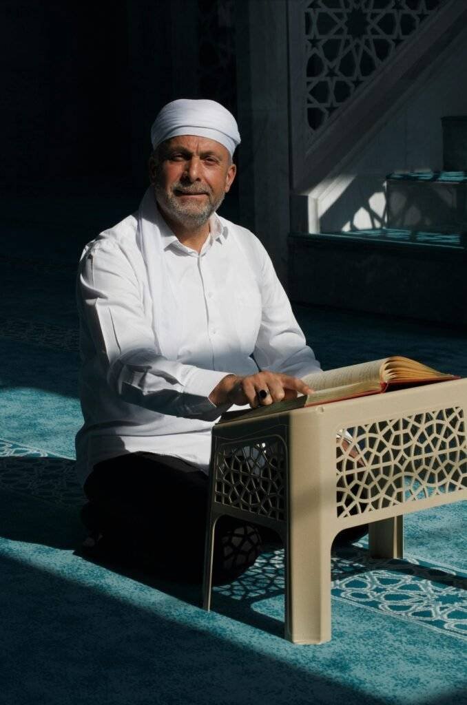 Middle-aged man in traditional attire reading a holy book in a serene mosque setting.
